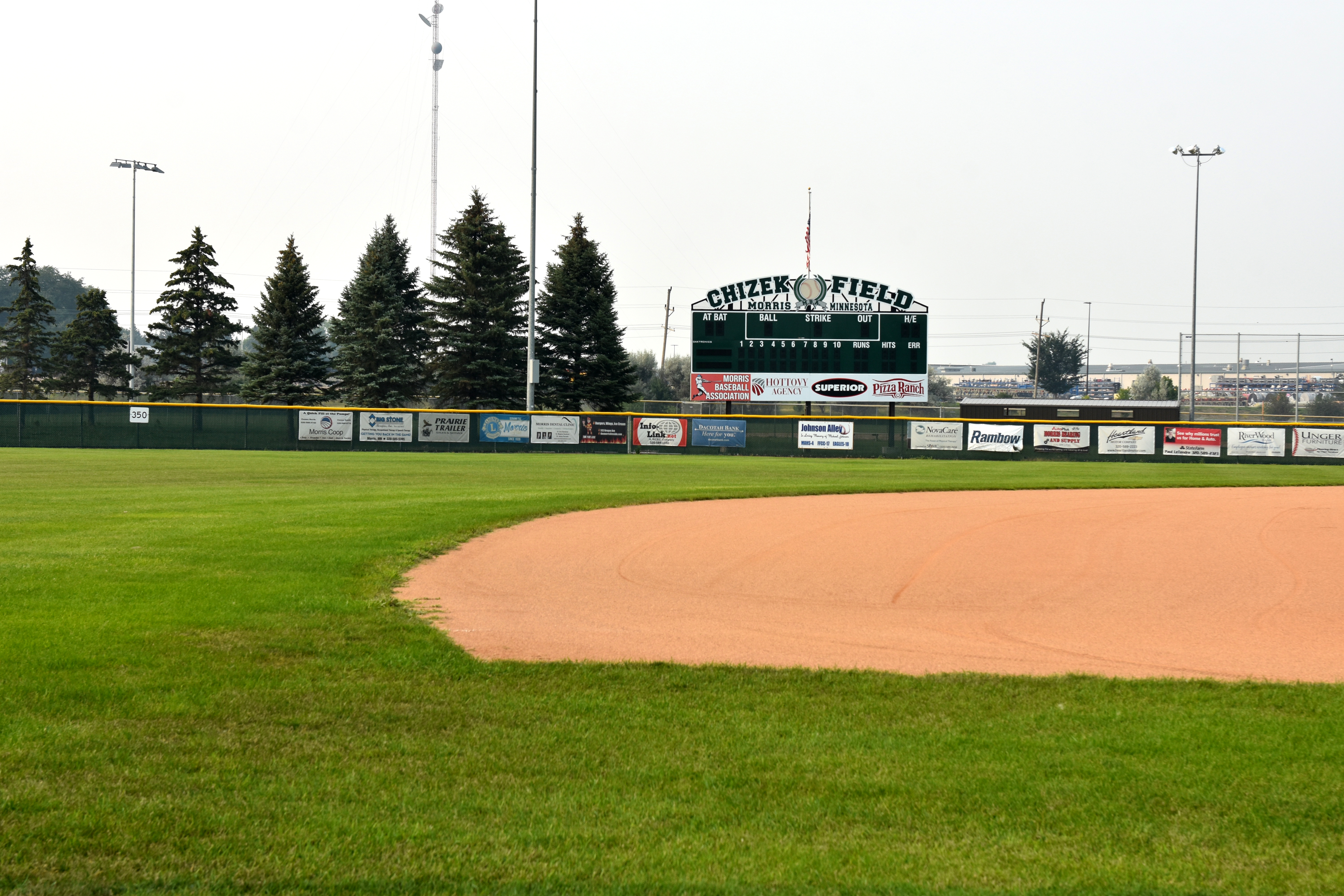 View across a baseball field with green outfield grass and an infield dirt curve leading toward the Chizek Field scoreboard and outfield fence lined with sponsor signs.